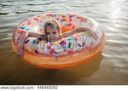 A Smiling Tween Girl Swims With A Big Donut Shaped Inflatable Ring On A Lake On A Hot Summer Day, Ha
