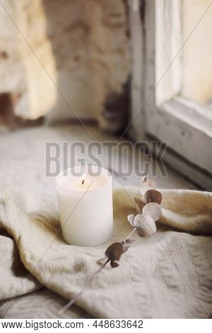 Close Up Of Burning Candle On Rustic Style Windowsill With Knitted Scarf And Dry Twig Lying Next, At