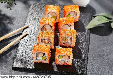Maki sushi on dark stone table. California maki with tobiko. Sushi roll with cheese,shrimp, avocado inside, maguro outside. Style concept japanese menu with black background, leaves and hard shadow