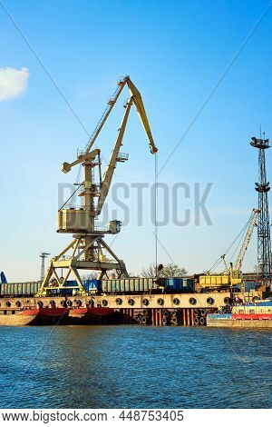 River Port Crane Loading Open-top Gondola Cars On Sunny Day. Empty River Drag Boats Or Barges By Pie
