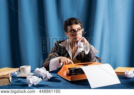 Thoughtful Newswoman Looking Away Near Crumpled Paper And Vintage Typewriter On Blue Background