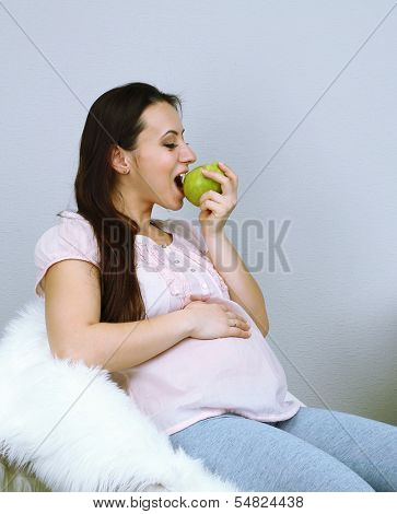 Young regnant woman sitting on armchair and eating apple on wall background