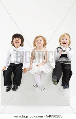 Little smiling girl and two boys sit on white big cubes on white background.