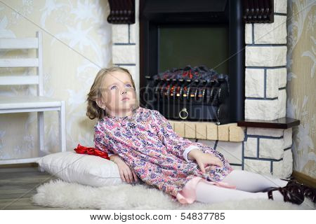 Little cute girl lies on white fur near fireplace in cosy room and looks up. 