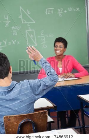 Rear view of teenage schoolboy raising hand while teacher looking at him in classroom