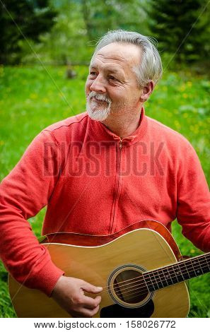 Portrait of a handsome senior man in bright sweater looking aside and plays acoustic guitar outdoor. Smiling happy senior man with gray hair and beard. Outdoors. Vertical image.