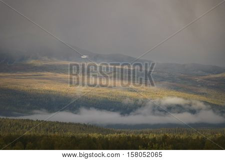 Sunlight giving golden glow to the autumn colored forest while moody clouds shrouding the landscape