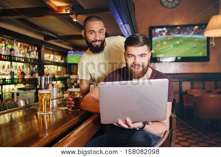 Two happy bearded young men drinking beer and using lapop in pub together