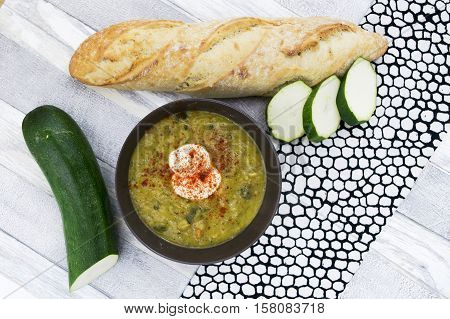 Courgette soup with sour cream in black bowl . Sliced zucchini and long fresh baguette on the white wooden background. Top view