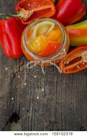 Bell pepper preserved in a glass jar with fresh peppers on old wooden background. Homemade marinated in oil red pepper. Glass jar with conserved roasted yellow and red paprika. Top view. Copy space.