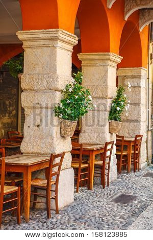 Wooden tables of comfortable outdoor cafe ristorante among ancient columns and archs at Verona streets. Flowers in flowerpots on poles. Front building exterior. Veneto region Italy