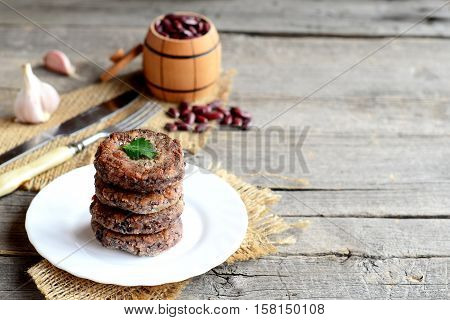 A stack of fried bean cutlets on a plate. Vegetarian cutlets cooked from red beans. Raw red beans in small barrel, garlic, fork, knife on old wooden background with copy space for text. Vintage style