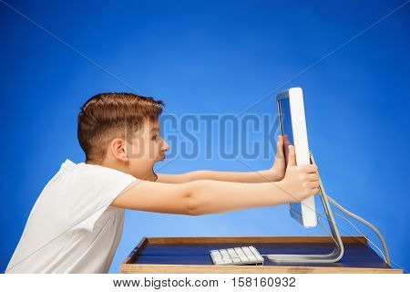 School-age boy sitting in front of the monitor laptop at blue studio backgroubd and screaming at the screen