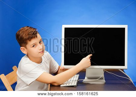School-age boy sitting with the monitor laptop and showing something at blue studio backgroubd