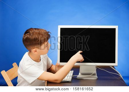 School-age boy sitting with the monitor laptop and showing something at blue studio backgroubd
