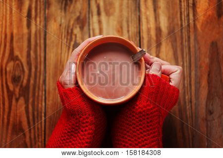 Woman holding cup of hot cacao or coffee on wooden background Closeup of hands in warm red sweaters. Top view