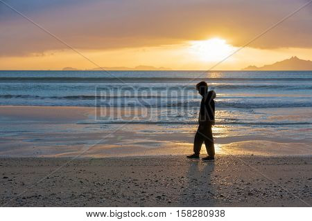Sunrise on beach Waipu Cove as boy silhouetted walks through glow of sun across water and beach