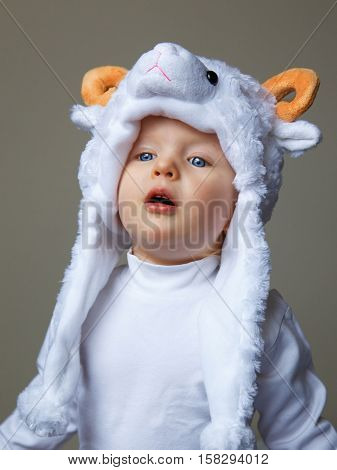 Cute adorable pretty baby with funny expression on her face making baa like a sheep wearing a sheep hat hood with yellow horns on top and white shirt on a light background New Year  concept studio