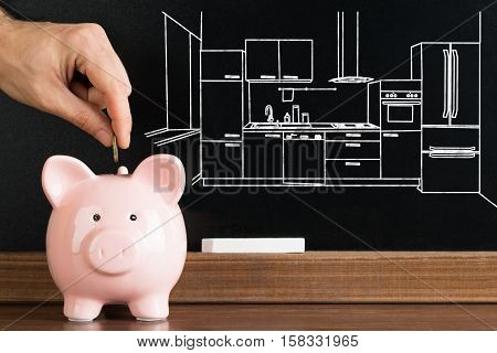 Close-up Of Person Inserting Coin In Piggybank With Kitchen Image Drawn On Blackboard In The Background