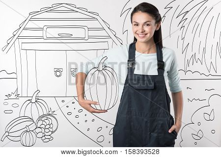 Nature products. Young pretty delighted farmer woman standing in the yard and smiling while holding a pumpkin.