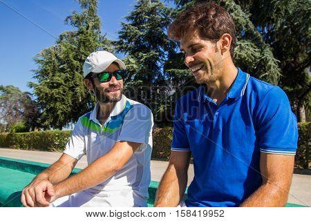 Two professional players hydrating after a hard game of tennis. They are friends and are sitting resting for the game. They are happy and laughing.