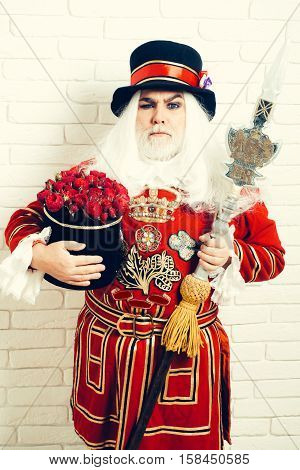 Frowning senior man beefeater yeomen warder or male royal guard bodyguard in red uniform with flowers and spear on white brick wall
