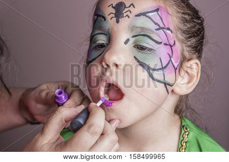 Make-up artist working with a little girl before halloween party. She is applying purple lip gloss