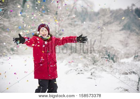 Happy kid boy with hands up with party confetti in winter park. Child having fun and Christmas celebration outdoors. Winter and holidays concept.