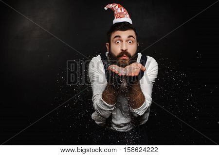 Young handsome man in Santa Claus hat blowing silver glitter over black background. Copy space.