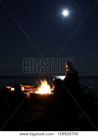 Woman and dog at a fire pit at seashore during full moon