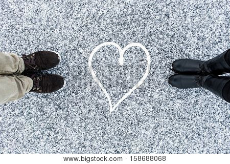 Male and Female boots standing at heart symbol on asphalt covered gritty snow surface. Rough snowy. Cold Winter. Top view. Relations concept