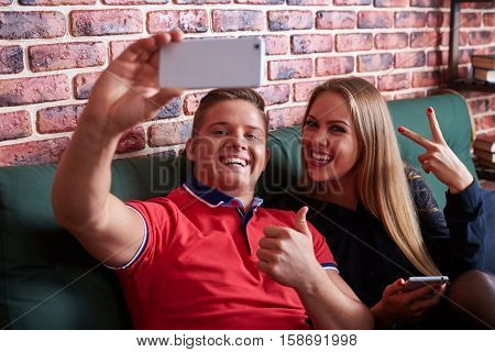 Close-up view of merry couple looking at the camera while making selfie. Extremely sweet happy couple sitting on the sofa and taking selfie via phone
