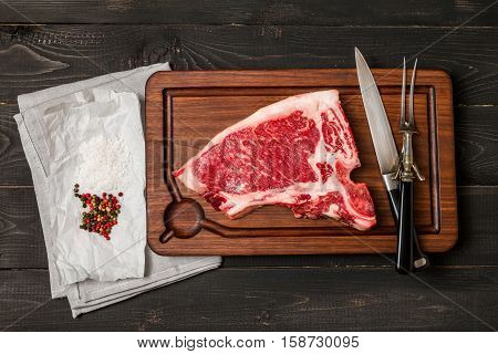 Raw fresh meat Club Steak, seasoning and meat fork and knife on dark wooden background, top view