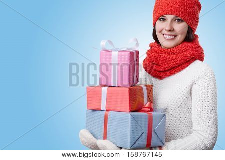 A woman wearing red hat with scarf white sweater and warm mittens being happy holding many presents in her hands. A woman with shining smile dreaming about New Year holidays on blue background