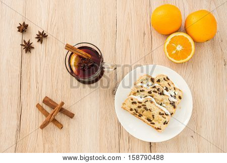 A of glass of Mulled Wine and slices of Christstollen surrounded by fresh oranges cinnamon sticks and star anise. Shot from directly above on light brown wooden background.