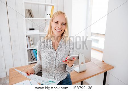 Cheeerful young business woman using mobile phone while standing at her workplace