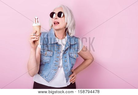 My passion. Emotional beautiful senior woman holding an icecrem and licking lips while standing against isolated pink background.