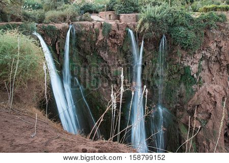 Red color of the mountain with several lines of waterfalls. Various bushes and trees growing on the mountain. Cascades Ouzoud Morocco.