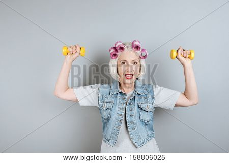 Sport is my life. Glad pretty woman smiling and exercising with dumbbells while standing against isolated gray background.