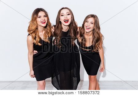 Three cheerful charming young women in black dresses standing and laughing over white background