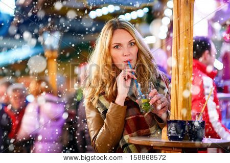 Beautiful young woman drinking hot punch, mulled wine on German Christmas market. Happy girl in winter clothes with lights on background. Family, tradition, holiday concept