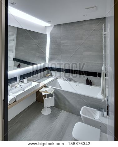 Bathroom in a modern style tiled with the white and gray tiles. There is a white sink with accessories, mirror, bath, towel holder, towel radiator, toilet and a bide, stand with a towel and a book.