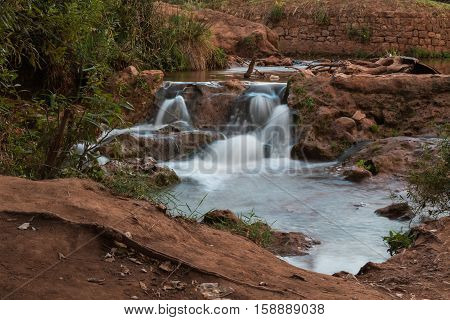 River in the city Cascades d Ouzoud surrounded by the trees and bushes of an oasis. Dark orange color of the soil. Terrain creating a small waterfall.
