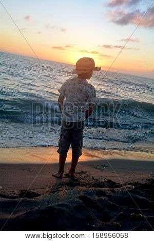 Small boy stands on the beach at the sundown, hands in the trouser pockets and looks aside