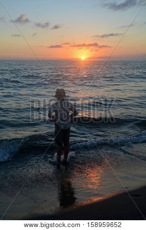 Small boy stands in the waves on the beach at the sundown and pulls up his trousers