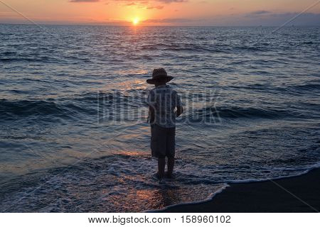 Small boy stands in the water on the beach at the sundown and looks in the water