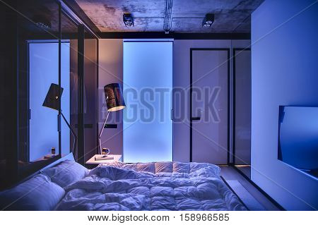 Low light bedroom in a loft style with light walls and concrete ceiling. There is a bed with pillows and blankets, table with book and clock, glowing lamp, TV. Light seeps through frosted glass door.