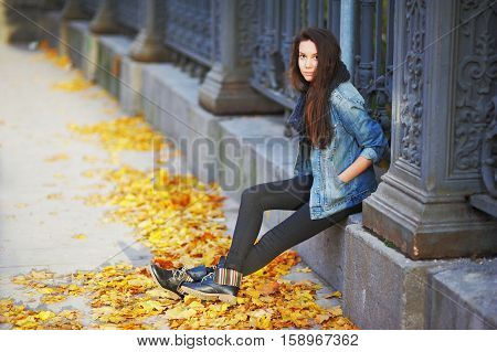 Young pretty woman sitting pensively on a stone slab a beautiful iron fence with his hands in the pockets of denim jacket around the yellow autumn leaves of maple side view.