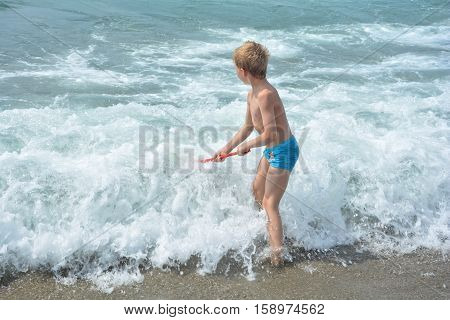 Small blond boy with blue swimming trunks stands with a fishing-net directly in the sea - wave
