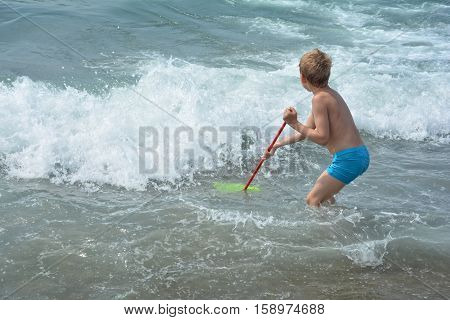 Small blond boy with blue swimming trunks stands with a fishing-net in the water and waits for the sea - wave